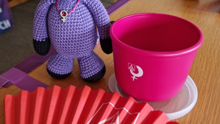 Das Bild zeigt Werbeartikel für das Seminar: einen pinken Becher, einen IG Metall Fächer und ein lilafarbenes gehäkeltes Teufelchen.
